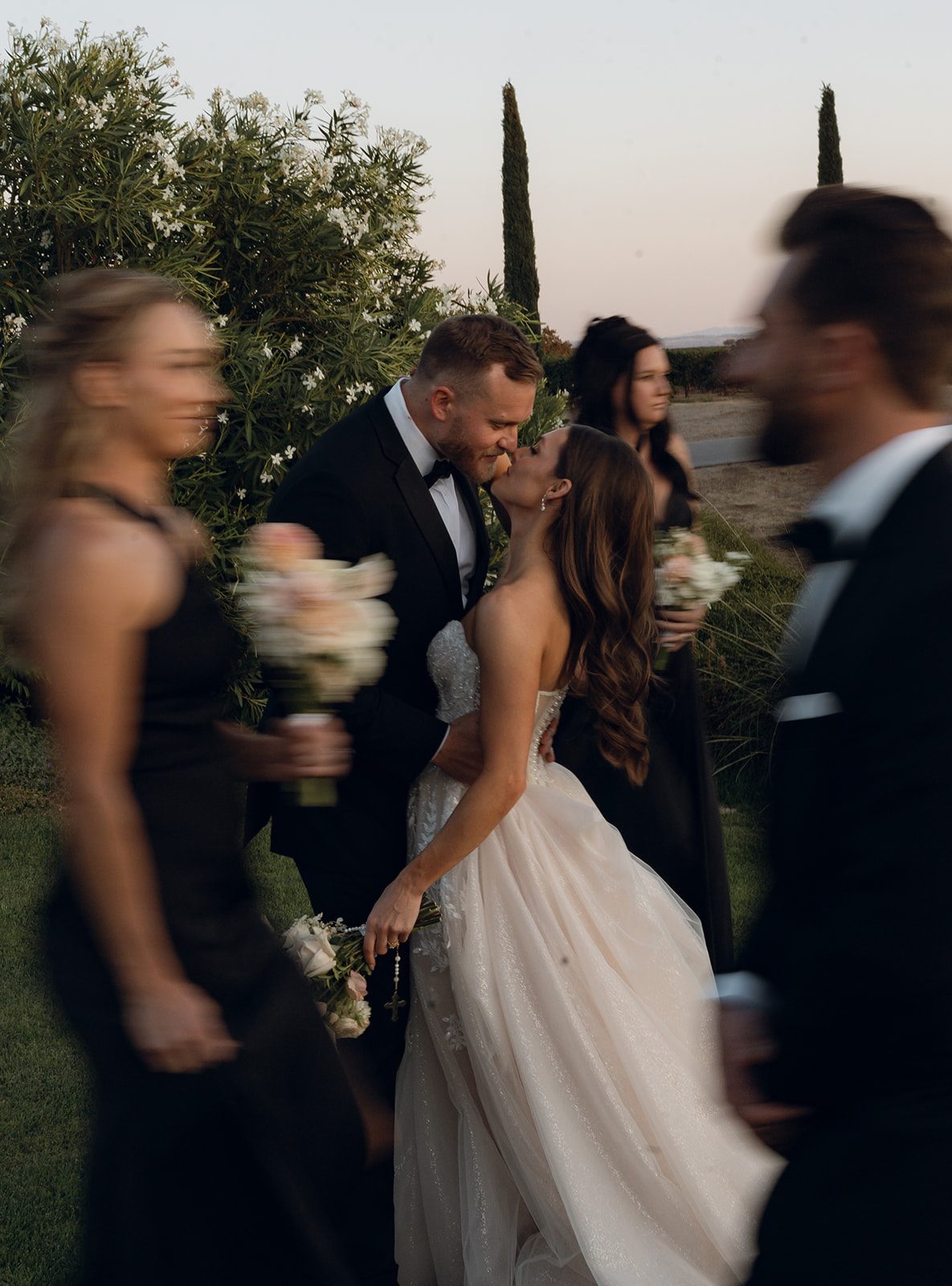 Bride and groom stealing a kiss surrounded by blurred wedding party members at dusk at a San Luis Obispo wedding venue