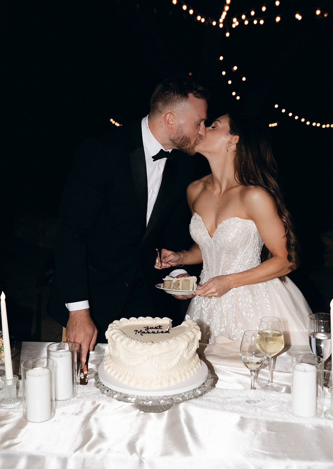 Bride and groom sharing a kiss while cutting their heart-shaped "Just Married" cake under string lights at an SLO wedding