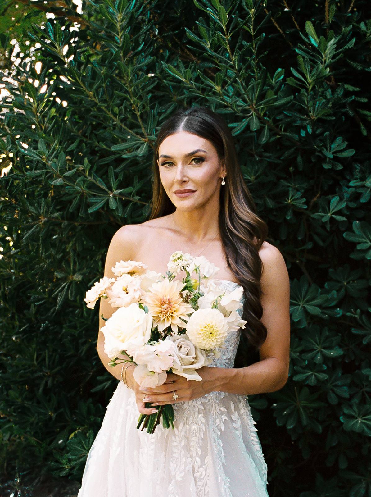 Bride in a strapless beaded gown holding a blush and ivory bouquet in front of a lush green hedge at Villa San Juliette