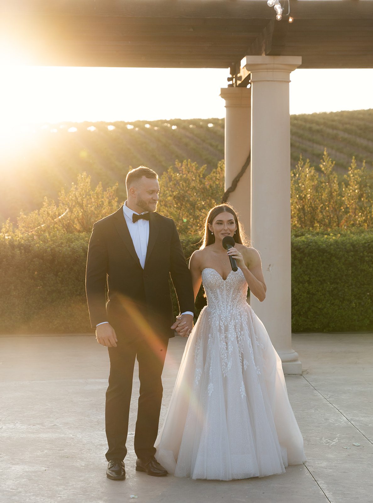 Bride speaking into a microphone while holding her groom's hand under the pergola with golden sunset light and vineyard views at Villa San Juliette