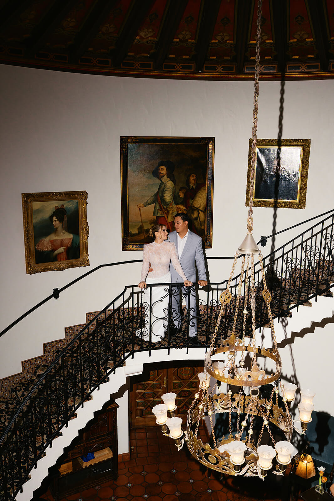 Couple standing on a curved staircase inside the Wrigley Mansion, surrounded by vintage artwork and a grand chandelier in this historic Arizona wedding venue.