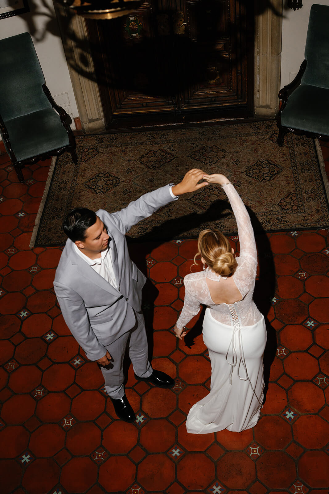 Overhead photo of a bride and groom dancing on red hexagonal tile flooring, with the groom in a light gray suit lifting the bride’s hand as she twirls. The bride wears a fitted gown with a lace-up back and long sleeves, her hair in a low bun. Vintage chairs and a patterned rug frame the scene, creating a warm, romantic atmosphere.