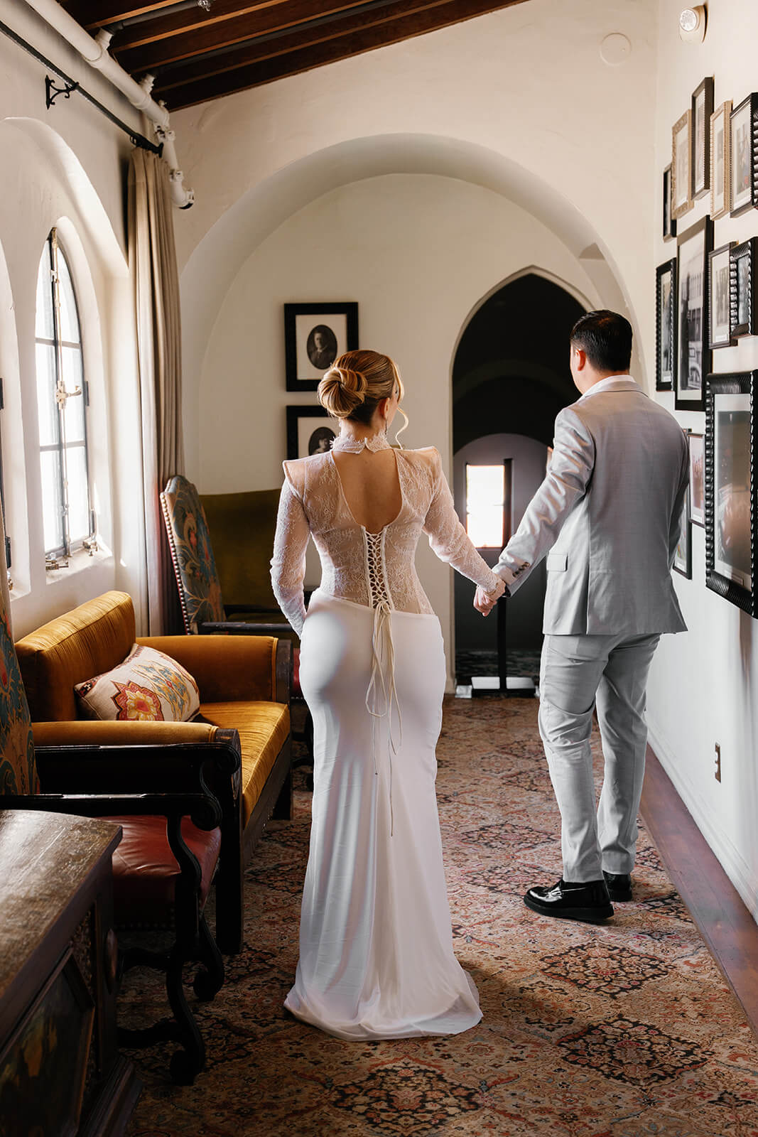 Bride and groom walking hand in hand through a warm, arched hallway, viewed from behind. The bride wears a fitted lace-back gown with long sleeves and an elegant updo, while the groom is dressed in a light gray suit. The space features rustic wood ceiling beams, a patterned rug, framed artwork on the walls, and soft natural light coming through a window, creating an intimate, timeless atmosphere.