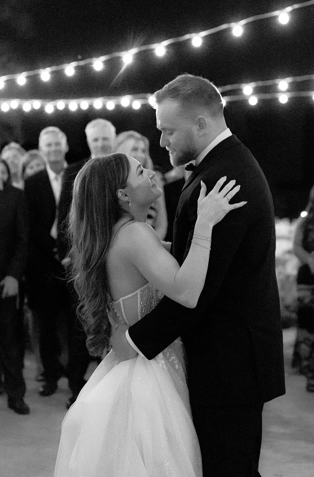 Black and white film photo of bride and groom sharing their first dance under string lights with guests looking on at a wedding venue in SLO county