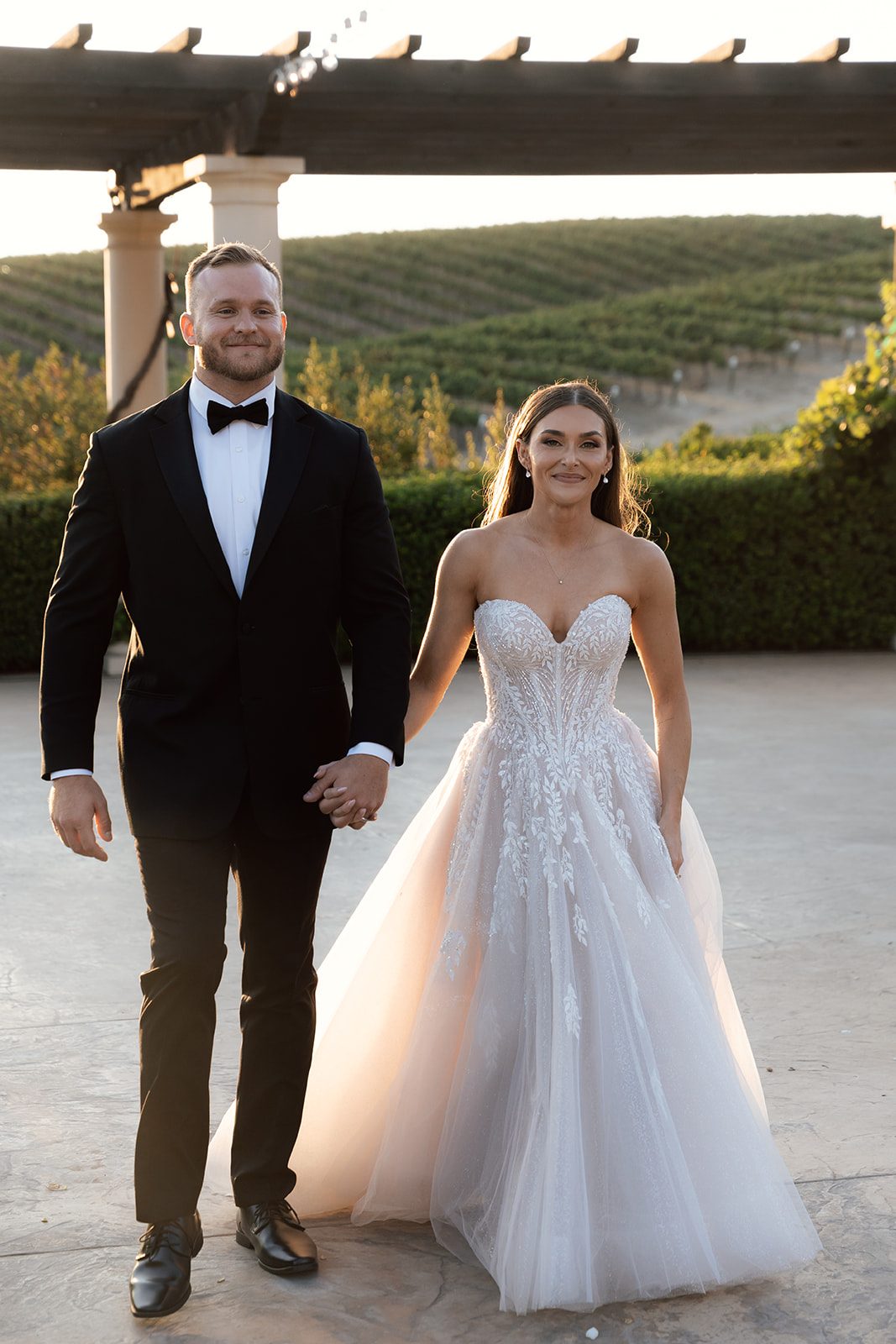 Bride and groom walking hand in hand toward the camera under the pergola with rolling vineyard hills glowing at golden hour at a wedding venue in SLO county