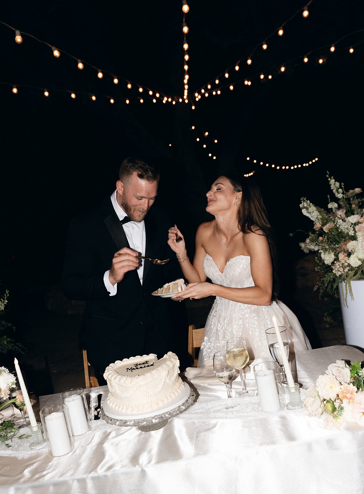 Bride and groom cutting their heart-shaped "Just Married" cake together under a canopy of string lights at an intimate wedding venue in California