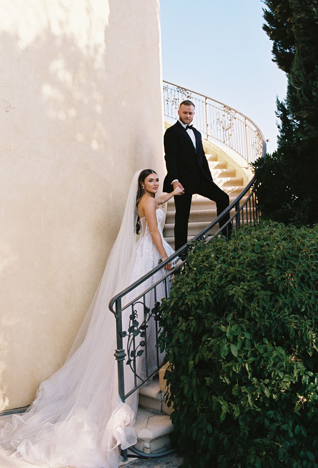 Bride and groom holding hands on the wrought iron spiral staircase with cypress trees and golden light at a San Luis Obispo wedding venue