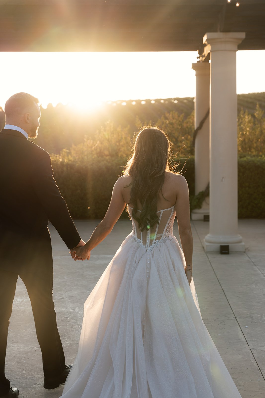 Bride and groom walking hand in hand toward the setting sun beneath the pergola at a wedding venue in SLO county