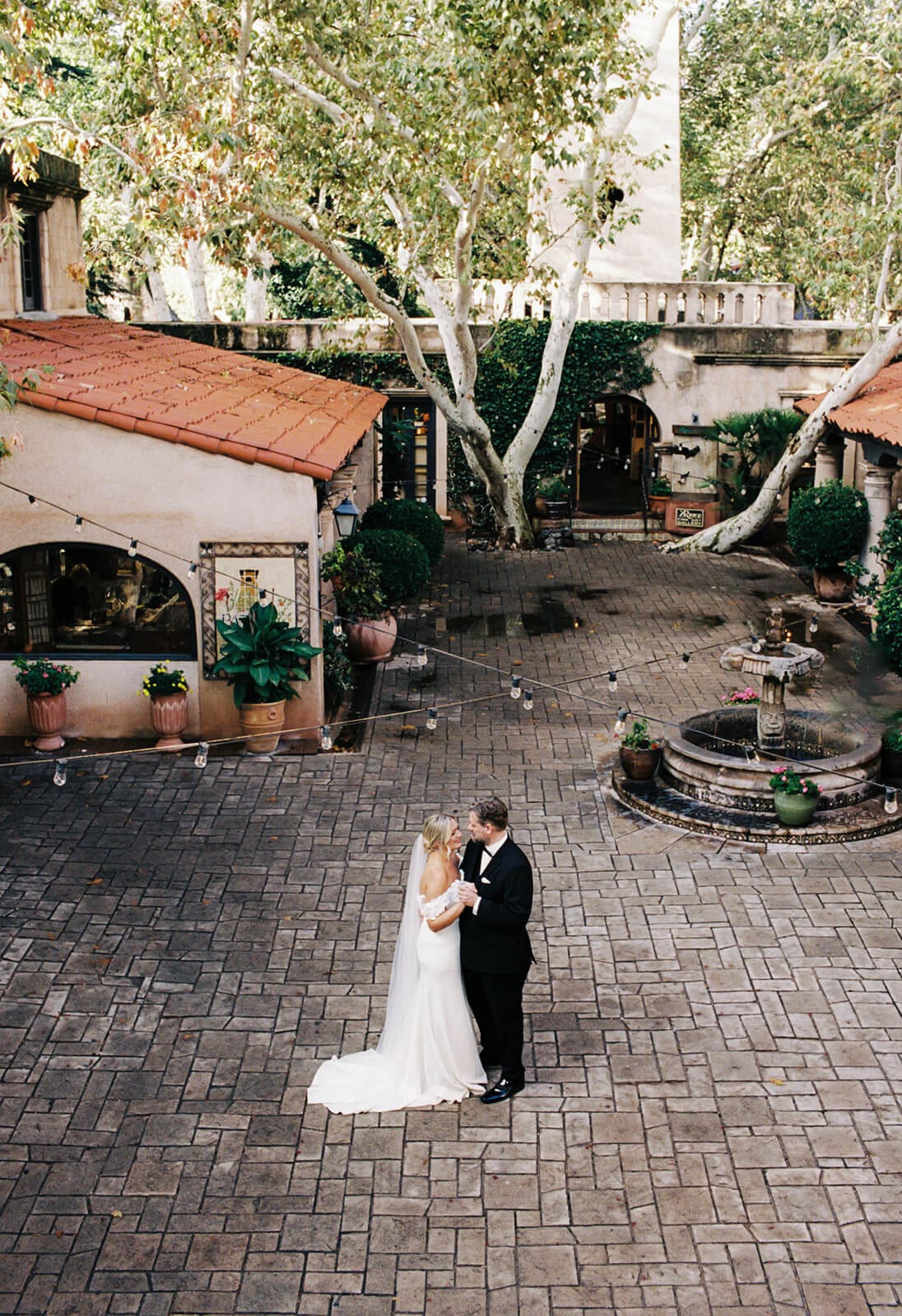 Bride and groom embracing in the center of a brick courtyard surrounded by trees, fountains, and Spanish-style buildings during a micro wedding at Tlaquepaque.