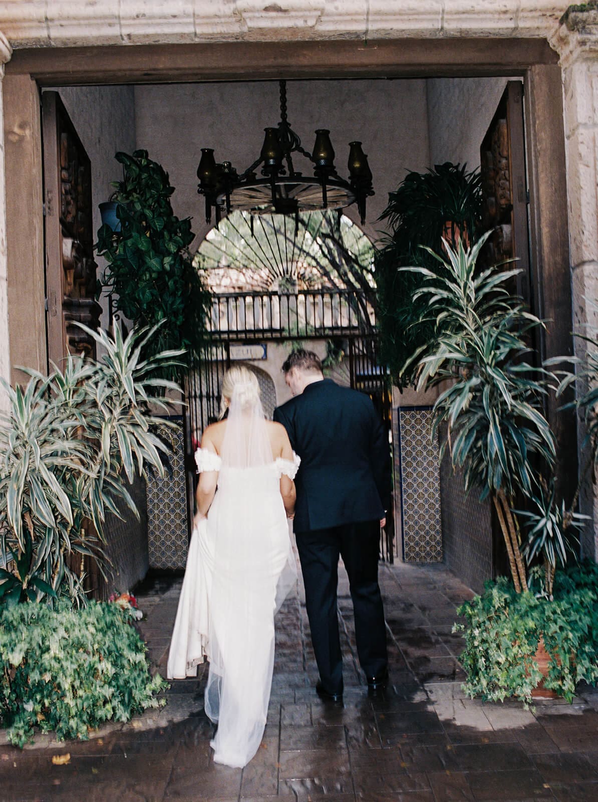Bride and groom walking through a large wooden doorway surrounded by tropical plants and Spanish-style architecture during a micro wedding at Tlaquepaque.