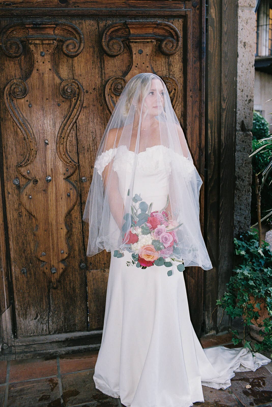 Bride standing in front of a carved wooden door holding a colorful bouquet during a micro wedding at Tlaquepaque.