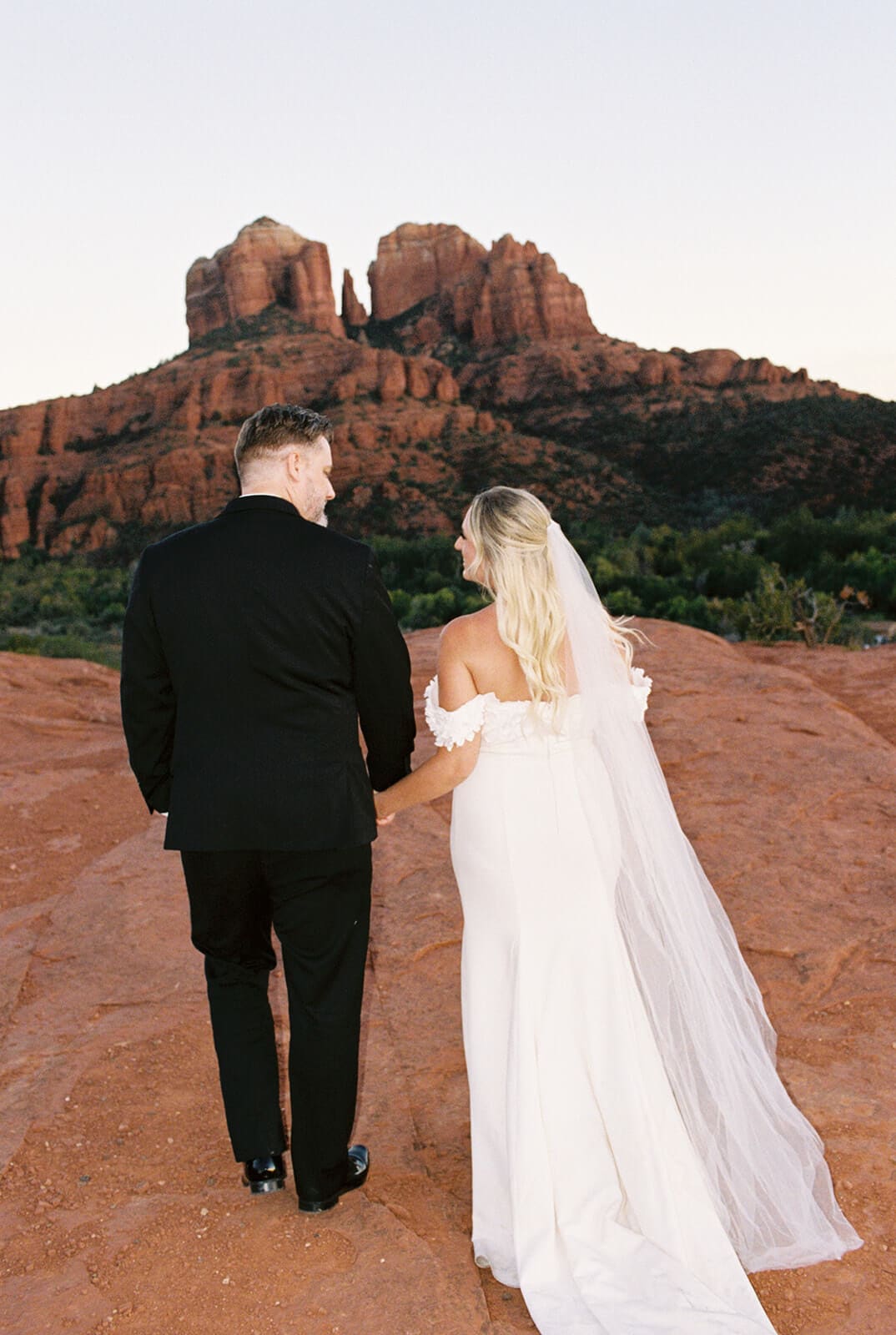 Bride and groom walking together on red rock cliffs during sunset in Sedona.