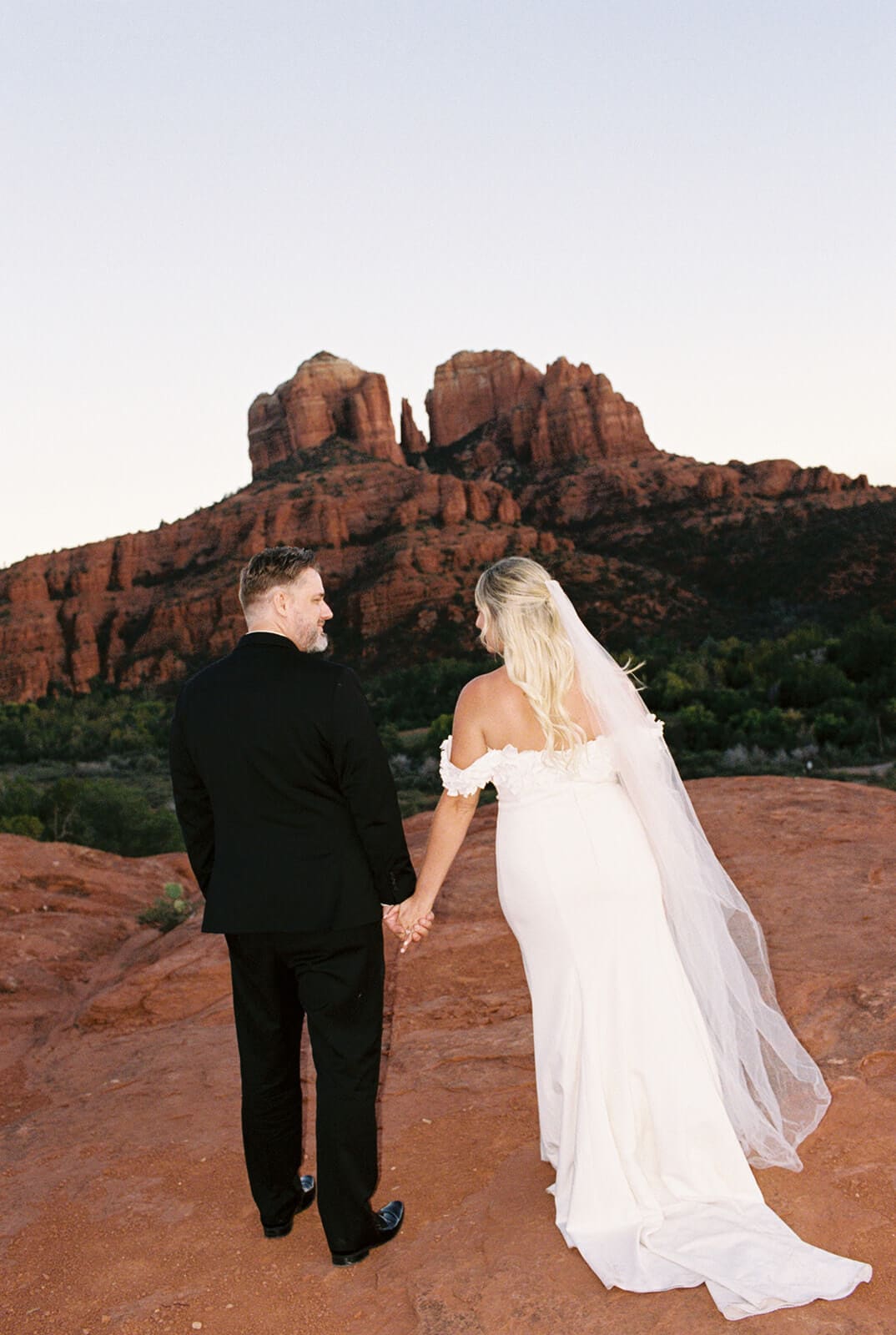 Bride and groom standing together on red rock cliffs with mountain formations in the background during sunset in Sedona.