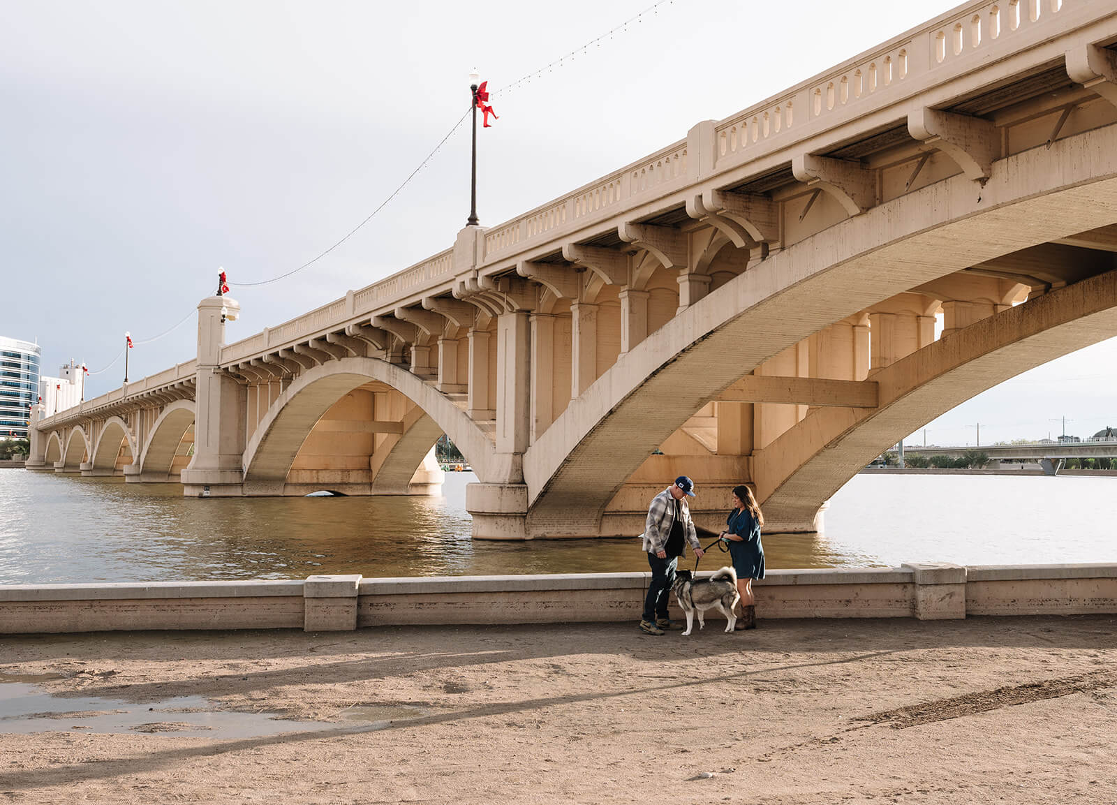 A couple stands with their husky dog along the waterfront at Tempe Town Lake, the arched bridge stretching across the water behind them.