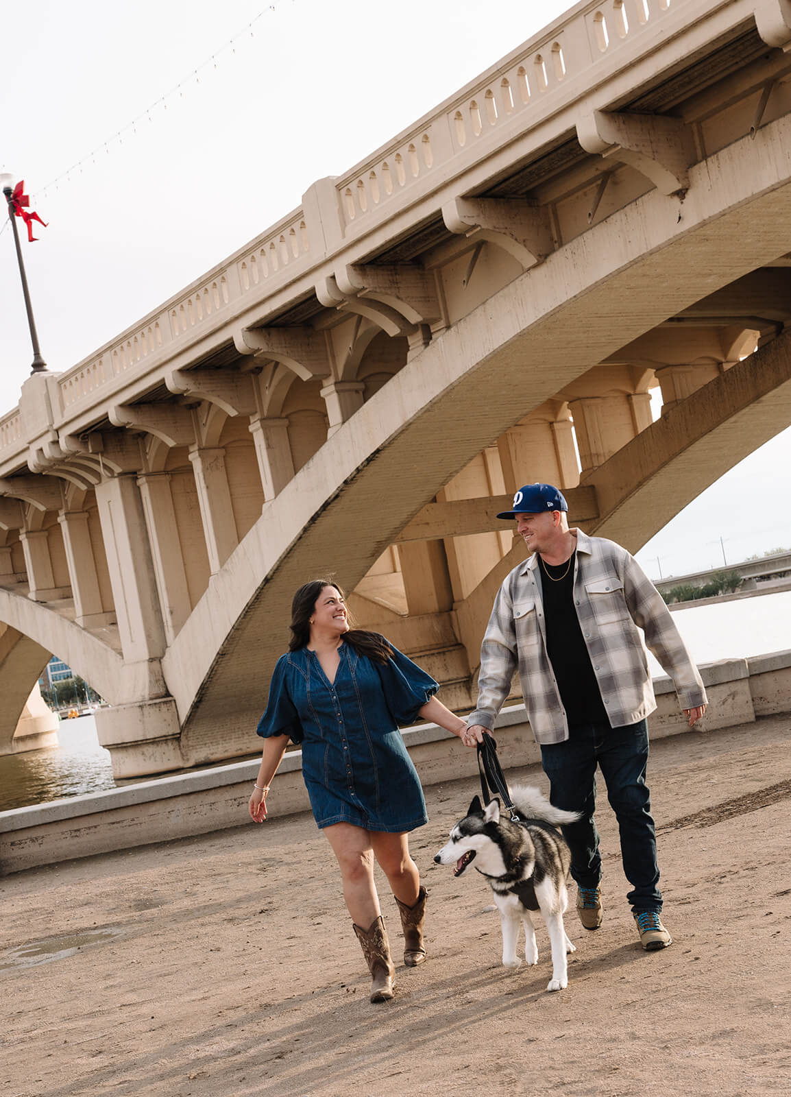 A couple walks along the sandy shore under the Tempe Town Lake bridge with their husky dog, the woman in a denim dress smiling up at her partner.
