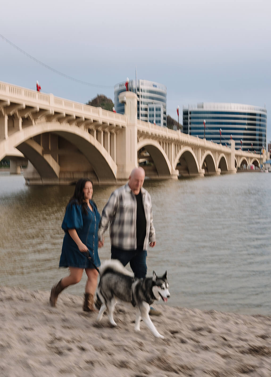 A motion-blurred photo of a couple walking along the sandy shoreline at Tempe Town Lake with their husky dog, the arched bridge and Tempe city skyline in the background.