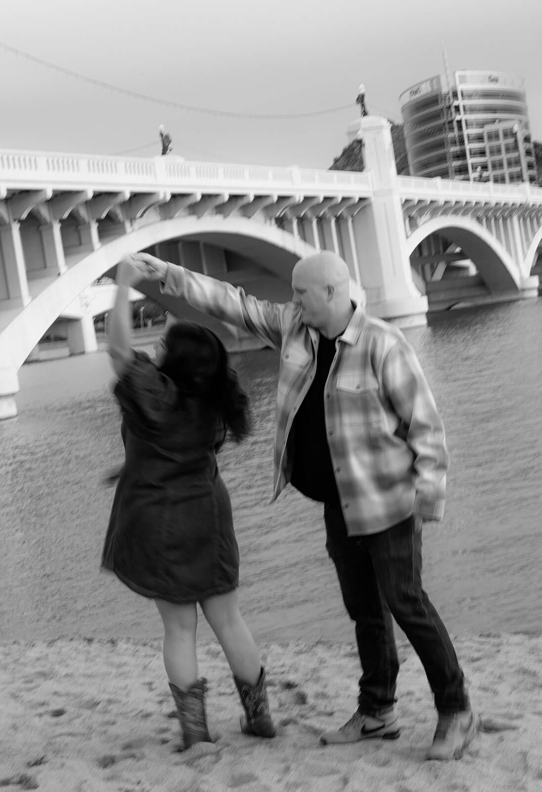 A black and white photo of a couple dancing and spinning on the sandy shore at Tempe Town Lake, the bridge and downtown Tempe skyline visible behind them.