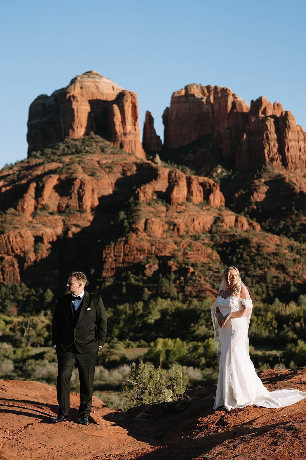 Bride and groom standing together on red rock cliffs with dramatic desert mountains in the background during sunset.