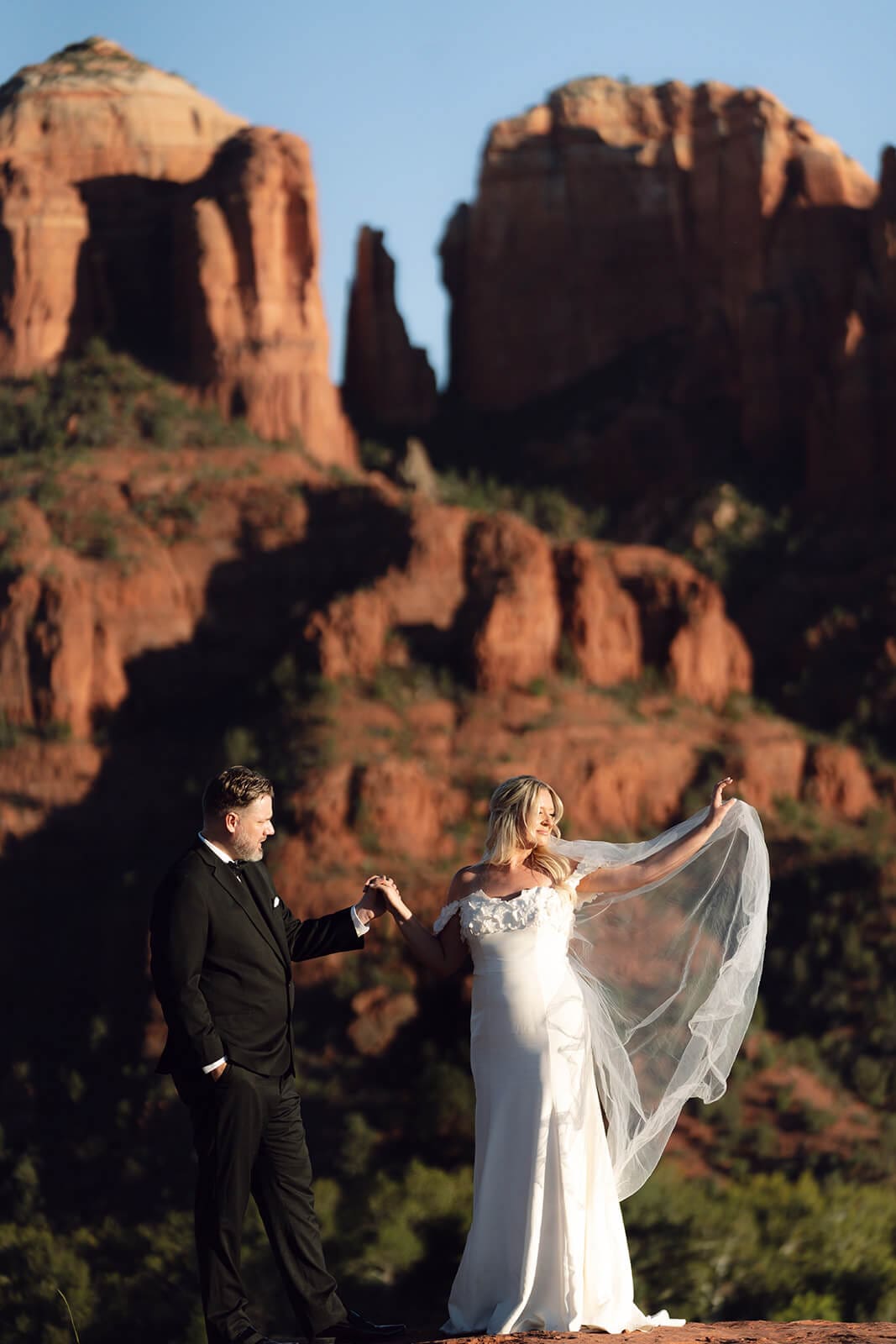Bride and groom holding hands on red rock cliffs with dramatic Sedona mountain views in the background.
