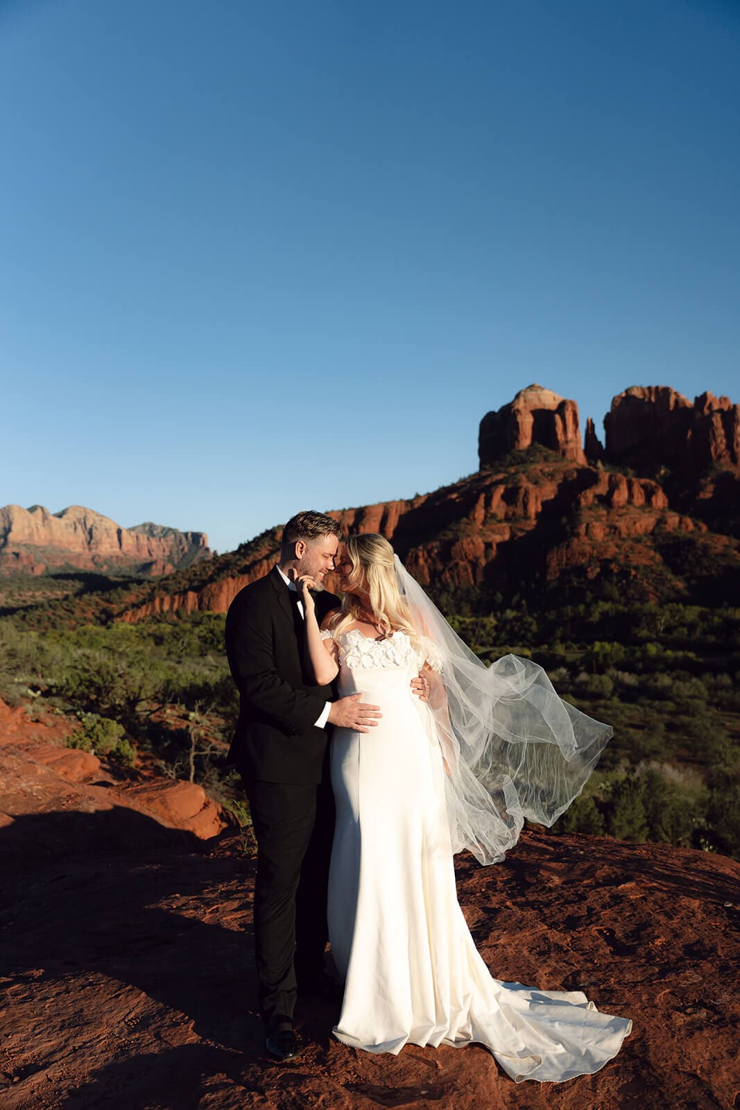 Bride and groom embracing on red rock cliffs during sunset with desert mountain formations behind them.