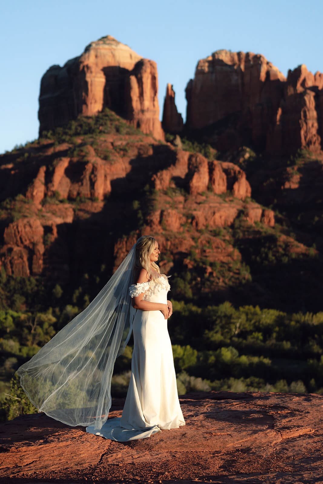 Bride standing alone on red rock cliffs with her veil flowing in the wind and mountain formations in the background.