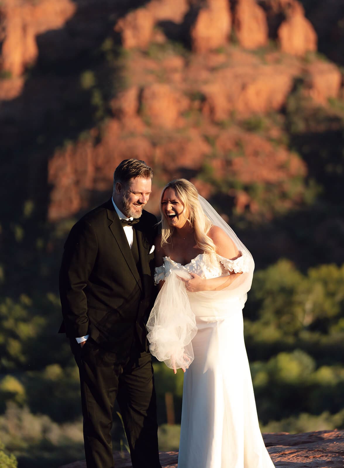 Bride and groom walking together on red rock cliffs during sunset with dramatic desert mountain views surrounding them.