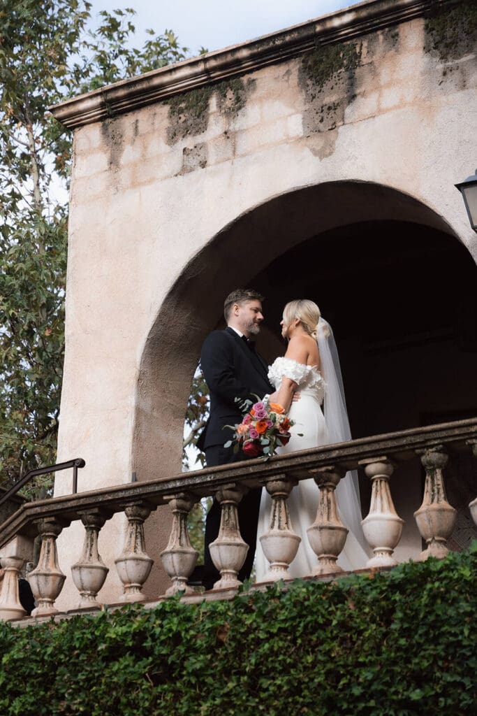 Bride and groom kissing on a stone balcony beneath a large arched opening at a historic venue.