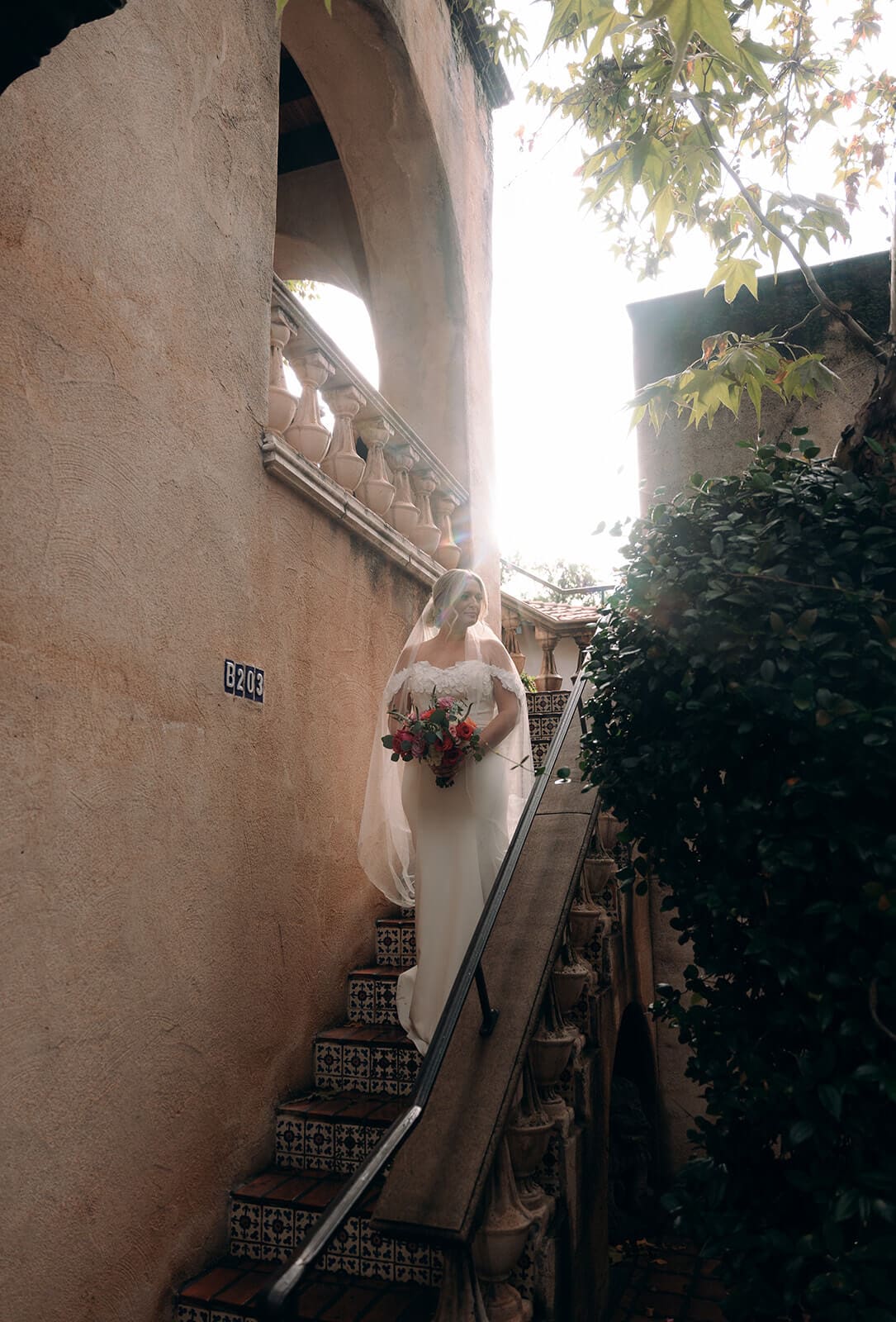 Bride walking up a narrow staircase beside a stucco wall with sunlight streaming through the trees during a micro wedding at Tlaquepaque.