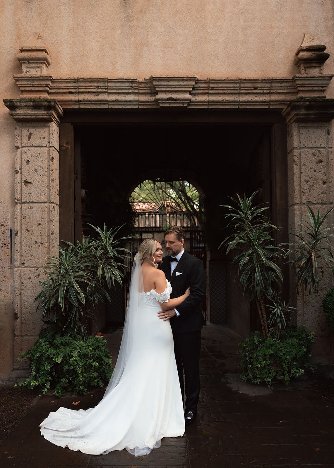 Bride and groom embracing beneath a large stone archway surrounded by greenery during a micro wedding at Tlaquepaque.