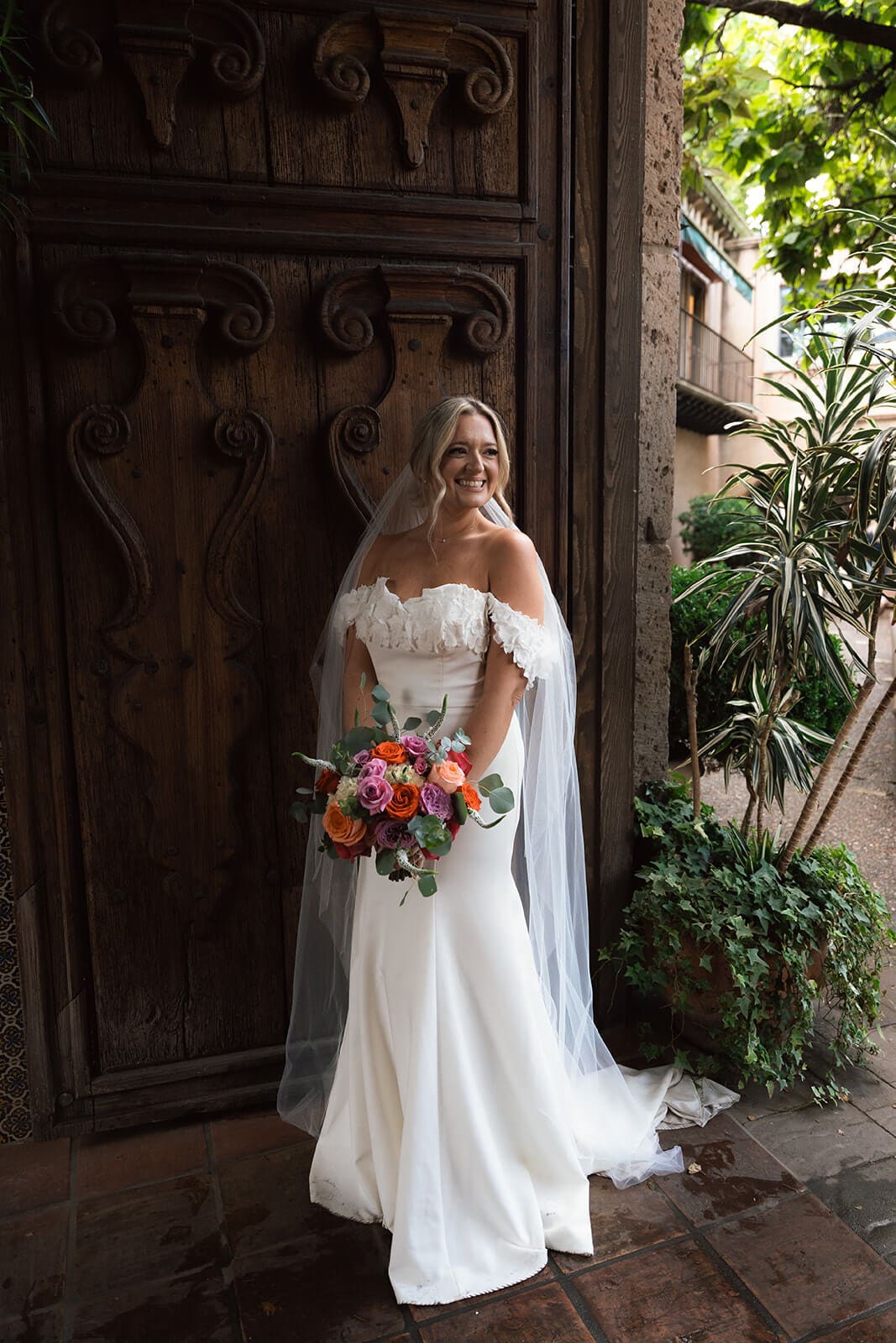 Bride standing in front of a carved wooden door holding a colorful bouquet during a micro wedding at Tlaquepaque.