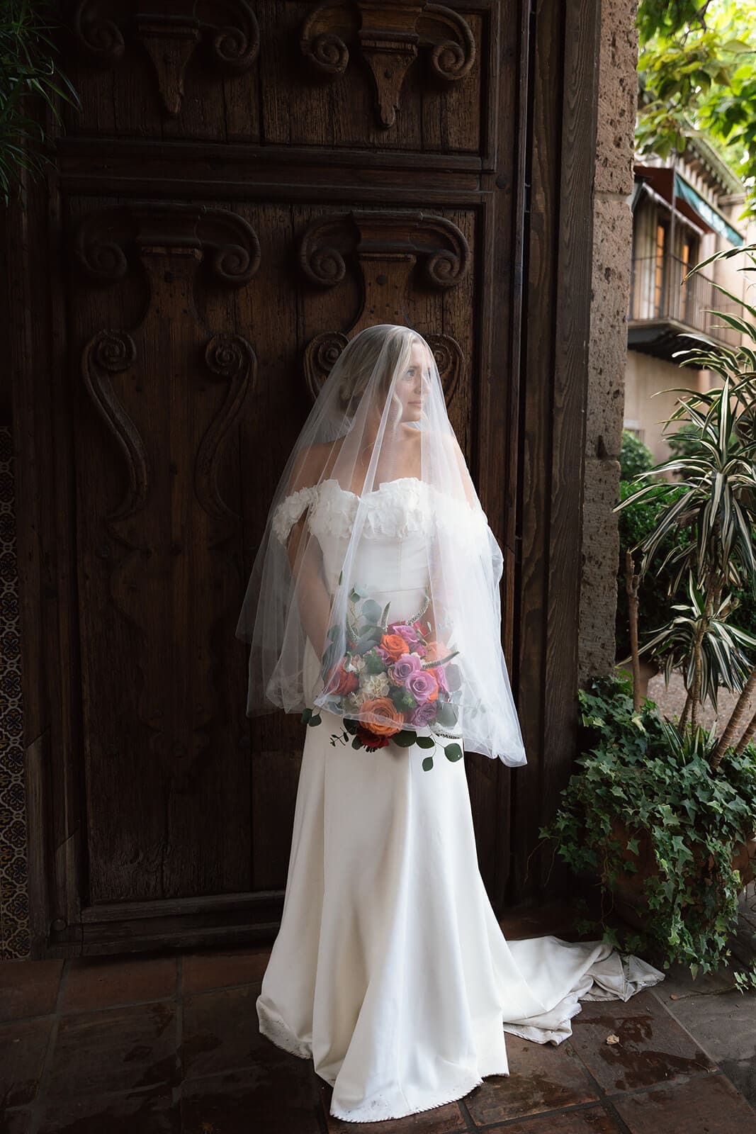 Bride standing in front of a large carved wooden door holding a colorful bouquet during a micro wedding at Tlaquepaque.