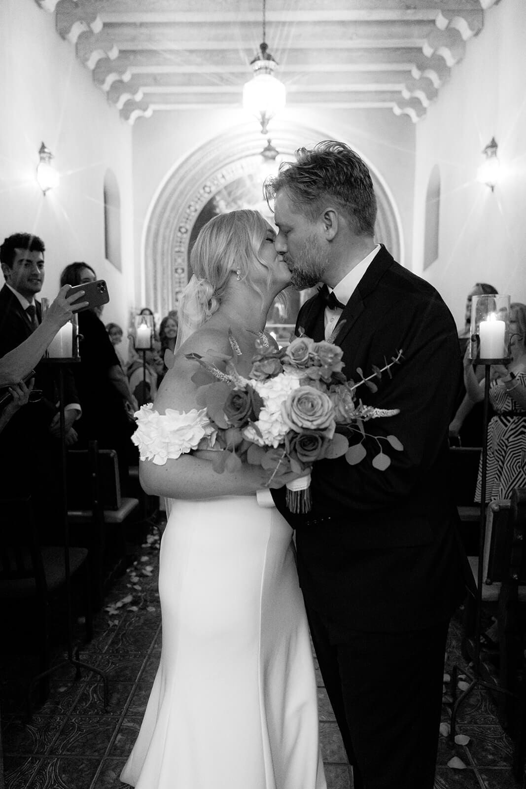 Black and white photo of a bride and groom kissing at the end of the aisle together after their ceremony inside a chapel during a micro wedding at Tlaquepaque.