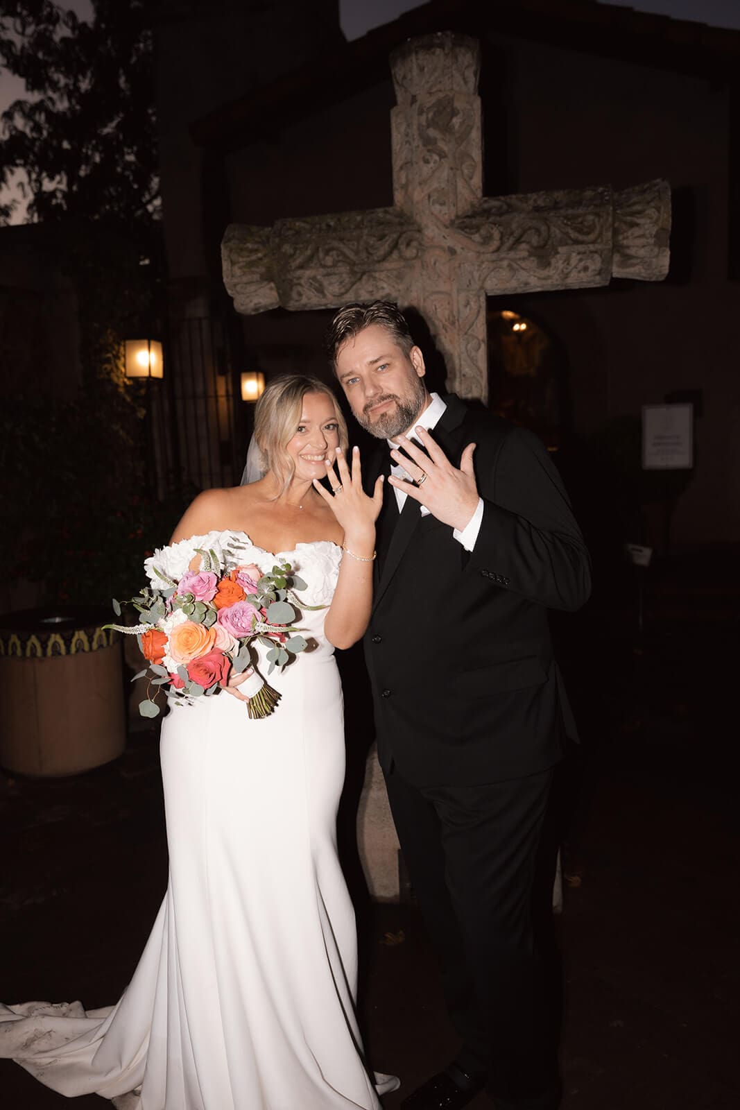 Bride and groom holding up their wedding rings and bouquet outside at night during their wedding reception.