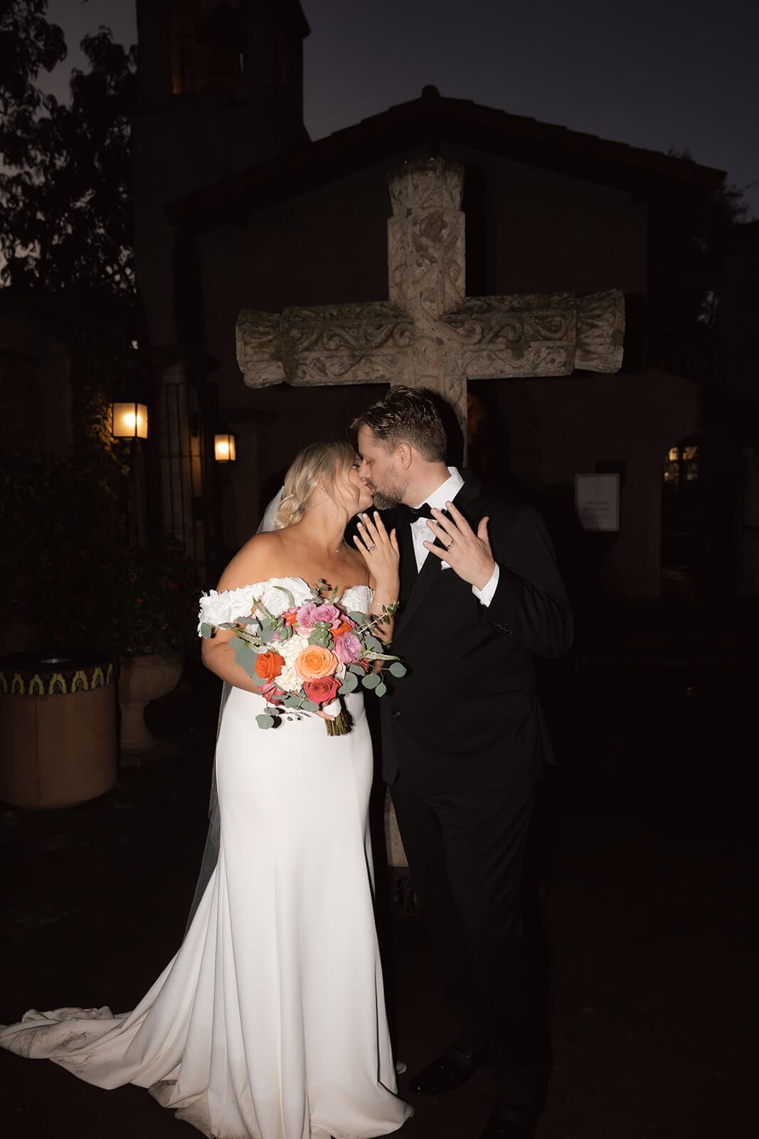 Bride and groom kissing while holding up their wedding rings and bouquet outside during an evening wedding reception.