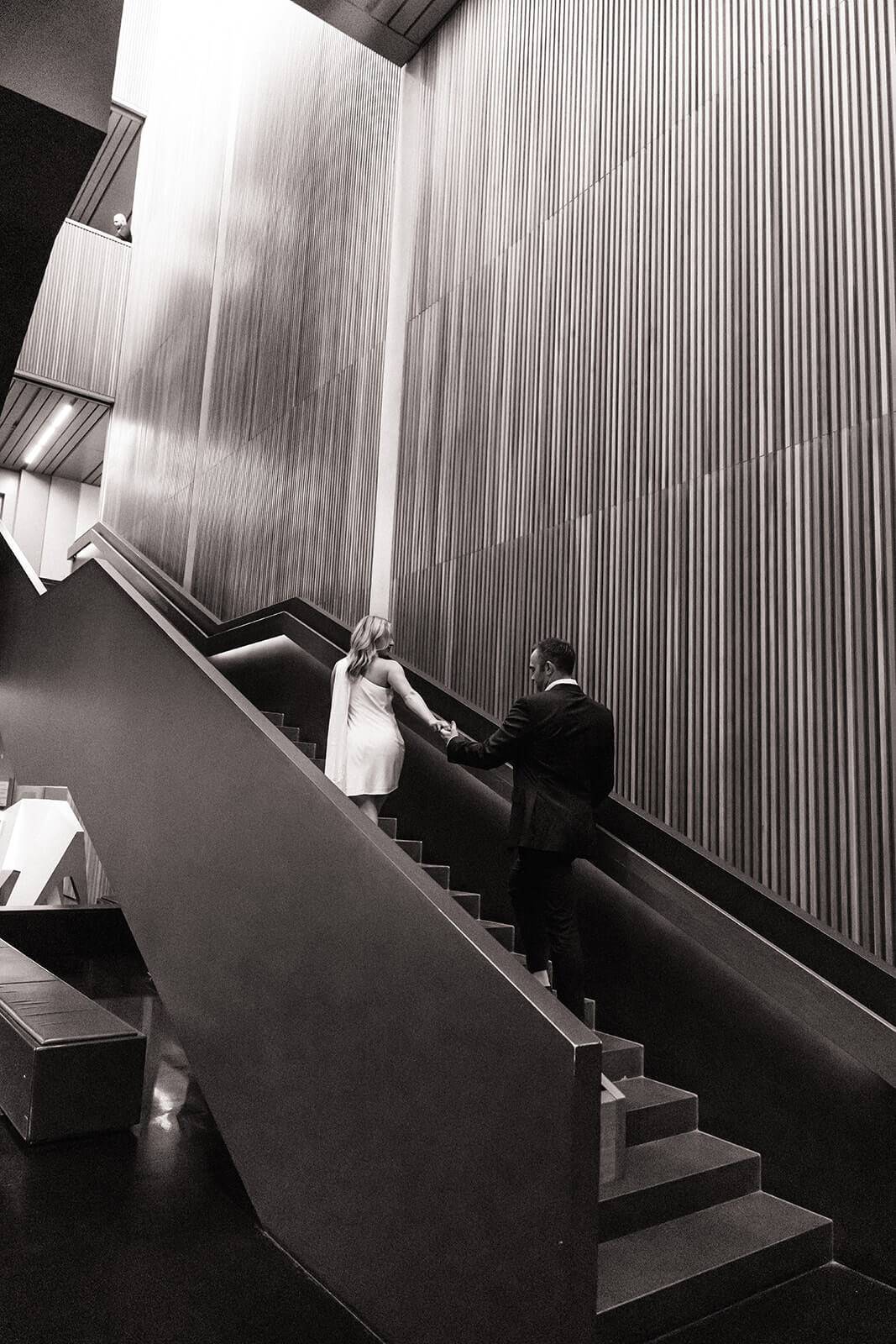 A couple holds hands while walking up a dramatic concrete staircase inside the Phoenix Art Museum, shot in black and white from above, one of the most editorial engagement photoshoot locations in Arizona.