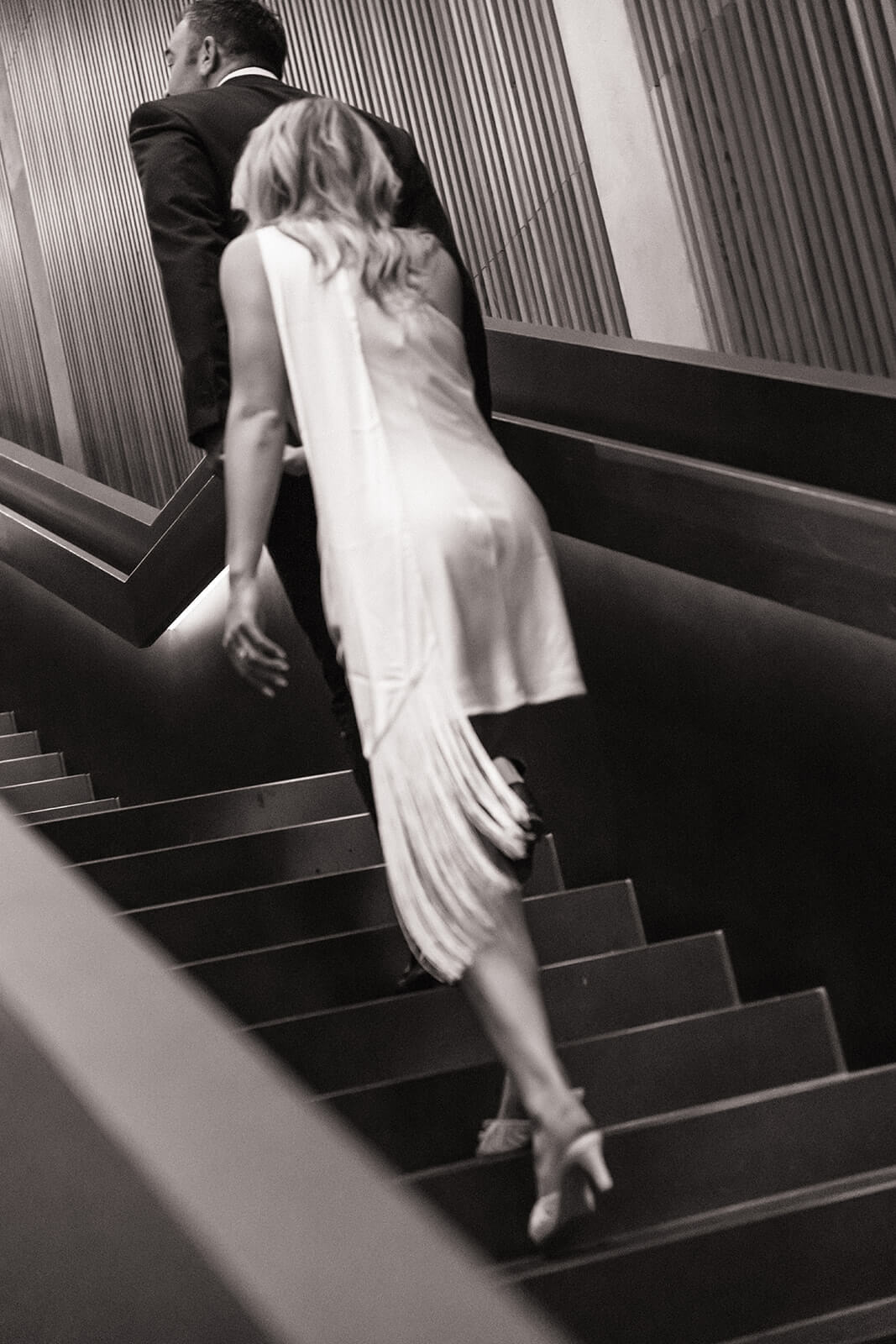 A close up black and white shot of a couple climbing the modern angular staircase inside the Phoenix Art Museum, the woman's white fringe dress trailing behind her.