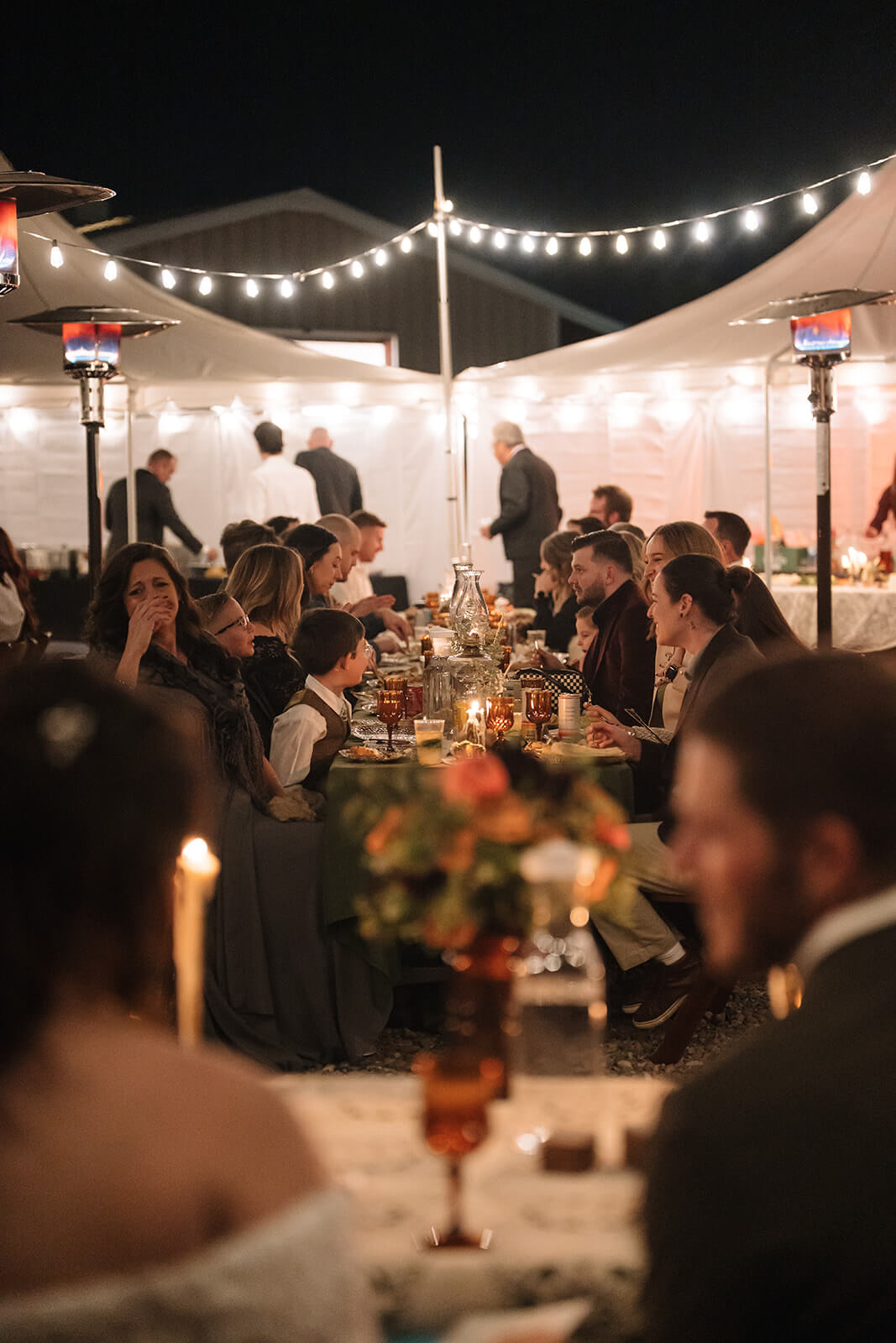 Intimate micro wedding reception dinner under string lights with guests gathered at a long candlelit table outdoors at night