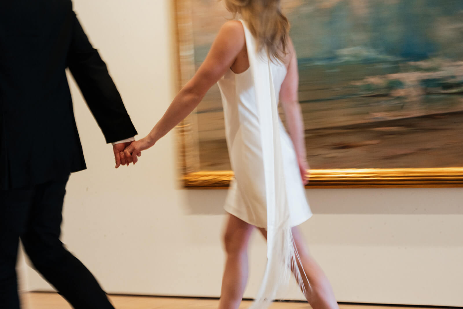 A motion-blurred photo of a couple walking hand in hand through a gallery inside the Phoenix Art Museum, a large gold-framed painting on the white wall behind them.