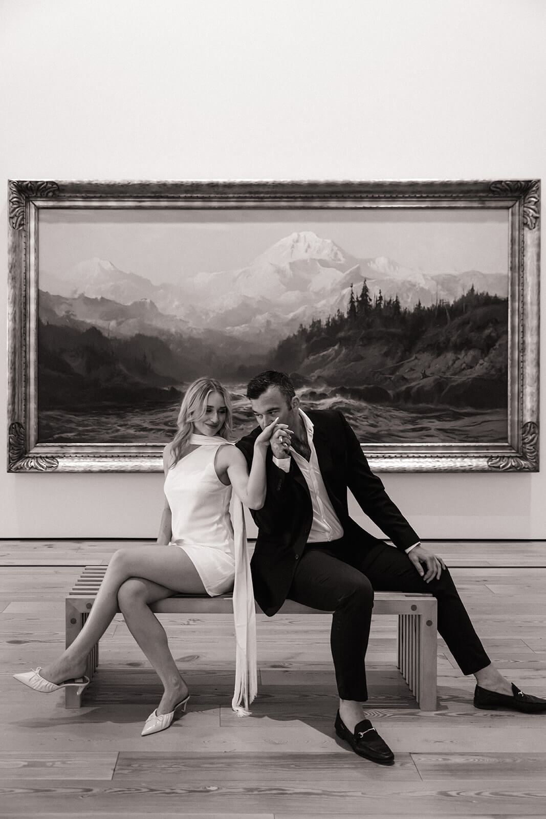 A black and white photo of a couple sitting close together on a gallery bench inside the Phoenix Art Museum, the man kissing the woman's hand in front of a large landscape painting.