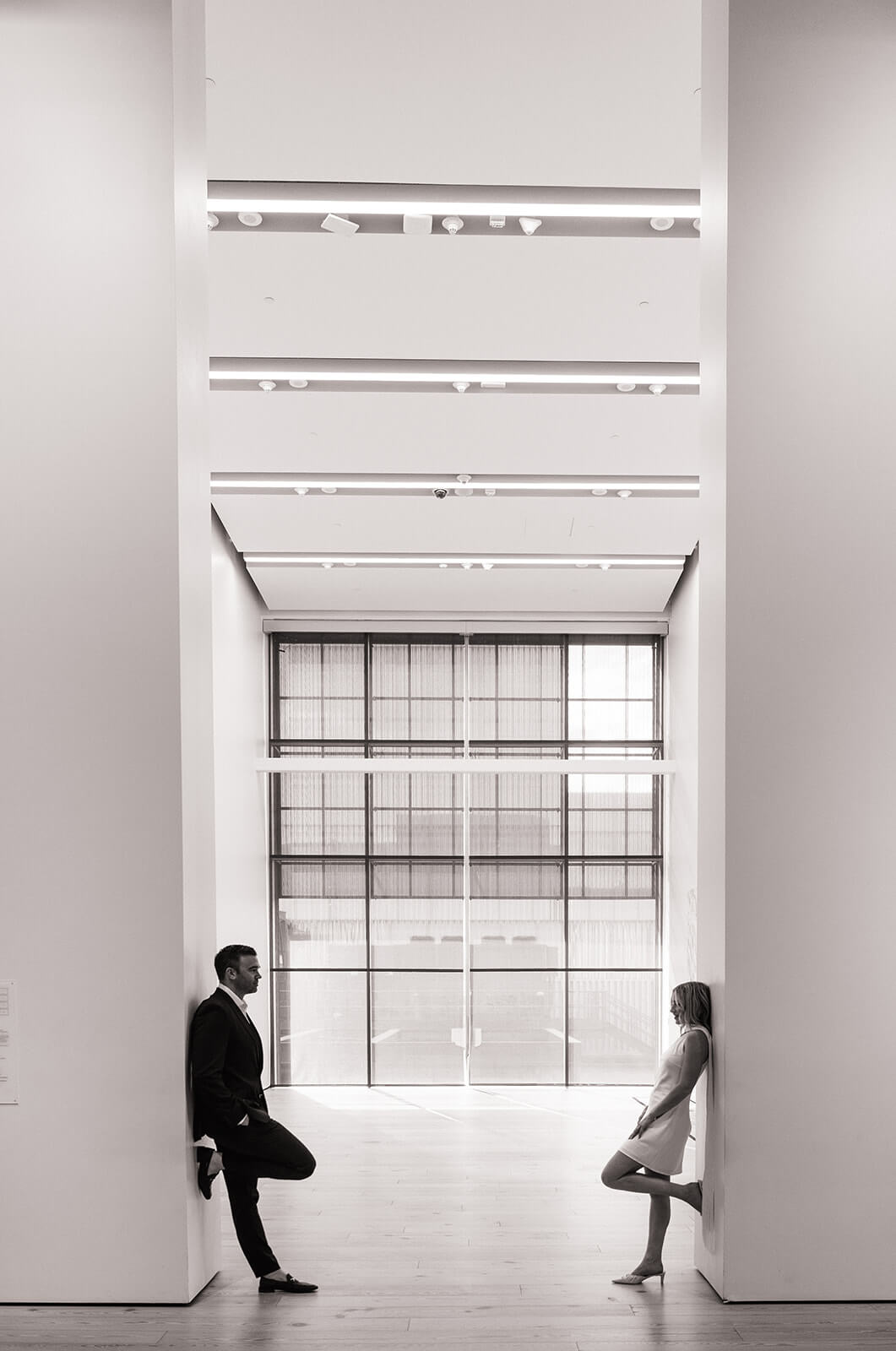 A black and white wide shot of a couple standing on opposite sides of a bright open corridor inside the Phoenix Art Museum, the tall ceilings and floor to ceiling windows making them look small against the architecture.