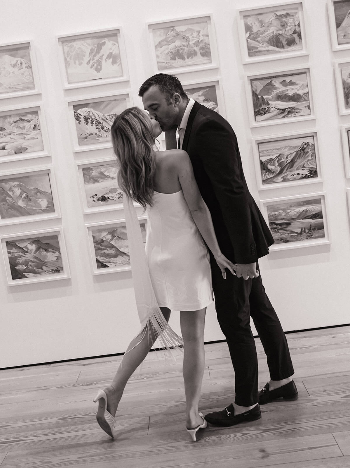 A couple shares a kiss inside the Phoenix Art Museum, shot in black and white with a grid of framed landscape artwork on the wall behind them.
