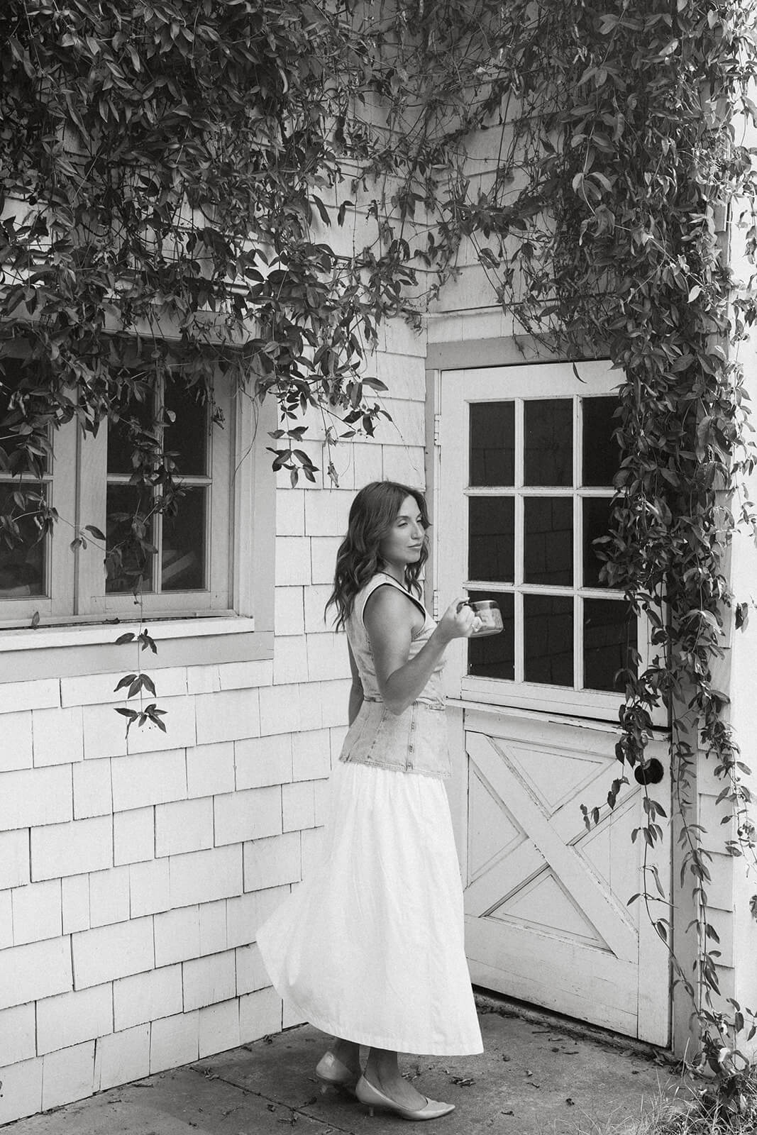 A black and white photo of a woman standing in front of an ivy covered white cottage door at The Bloomstead, looking back over her shoulder.