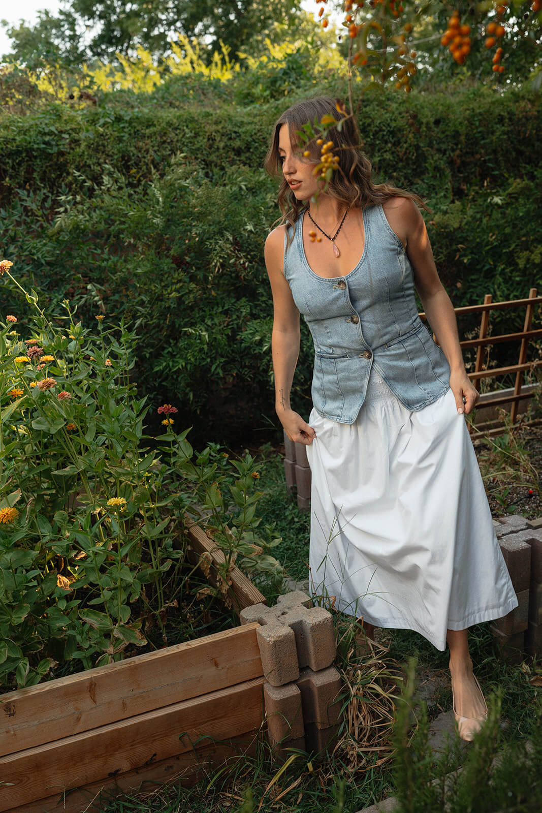 A woman in a denim vest and white skirt steps through a raised garden bed surrounded by lush greenery and orange blooms at The Bloomstead flower farm in Arizona.
