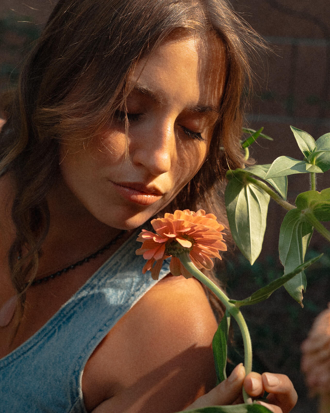 A close up portrait of a woman with her eyes closed, warm sunlight falling across her face as she holds an orange zinnia flower up to her chin at The Bloomstead flower farm.