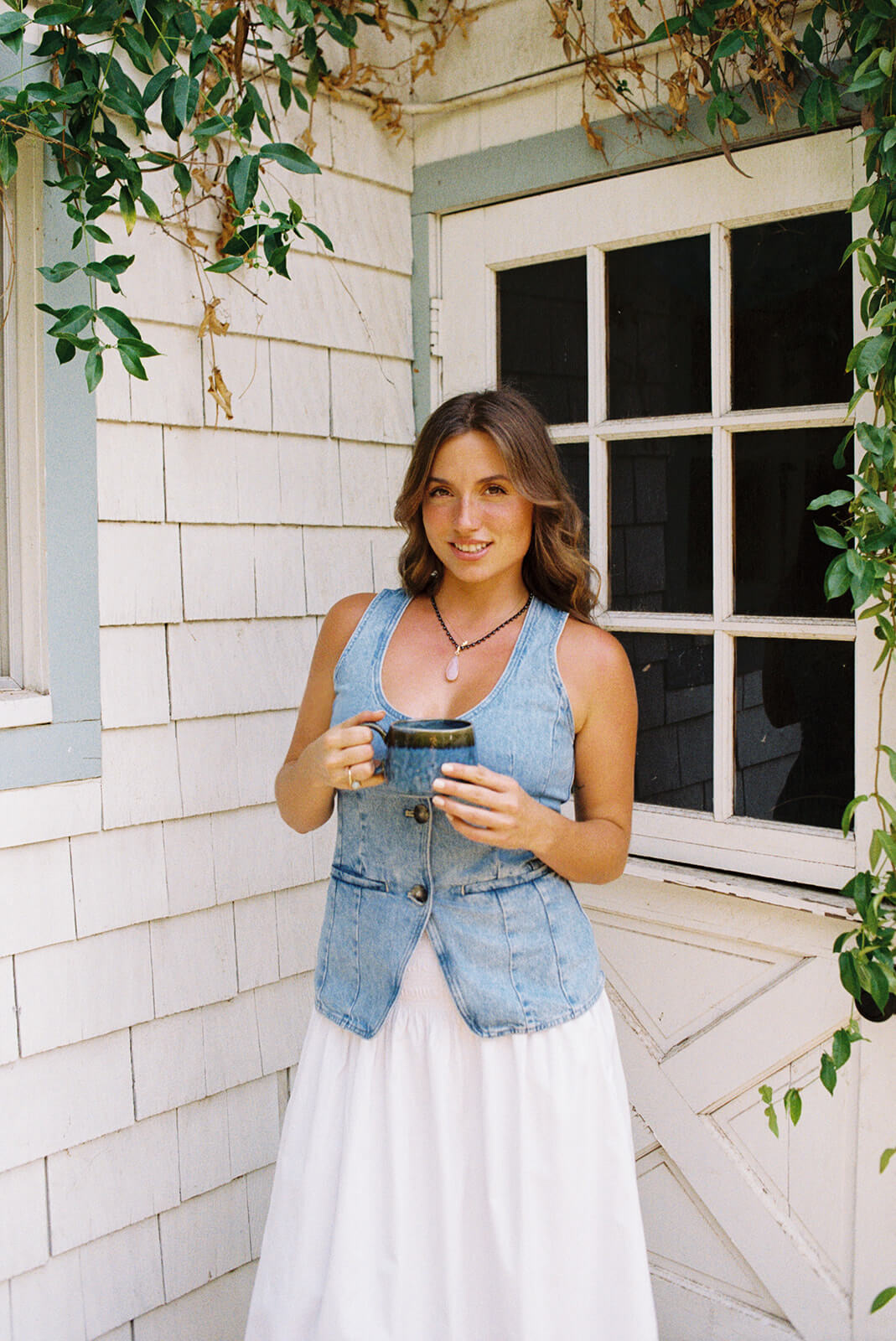 A woman in a denim vest and white skirt stands outside a white shingle cottage at The Bloomstead holding a coffee cup, ivy and greenery framing the window behind her.
