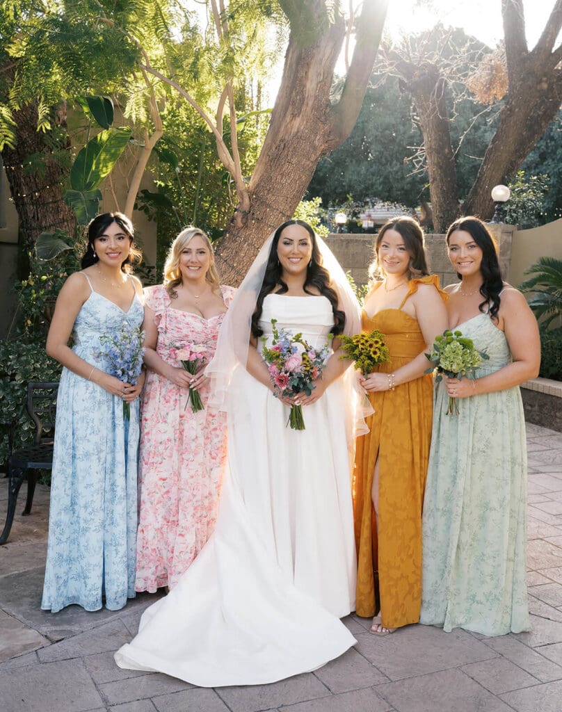 Bride posing outdoors with four bridesmaids wearing colorful floral and pastel dresses.