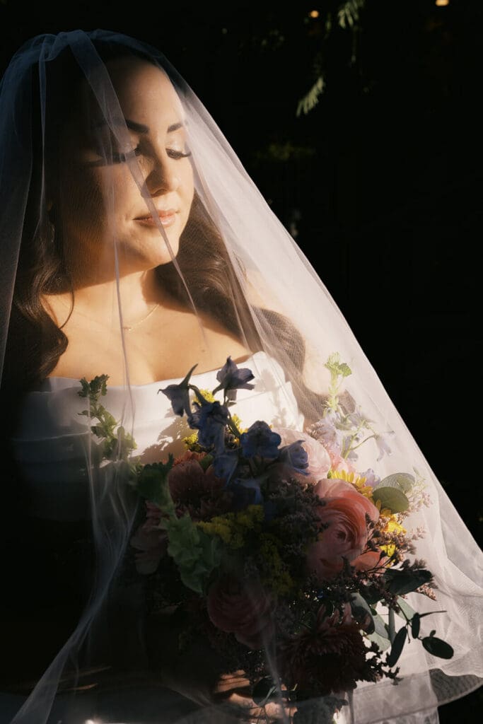 Close-up bridal portrait beneath a sheer veil with warm sunlight falling across her face and colorful bouquet.