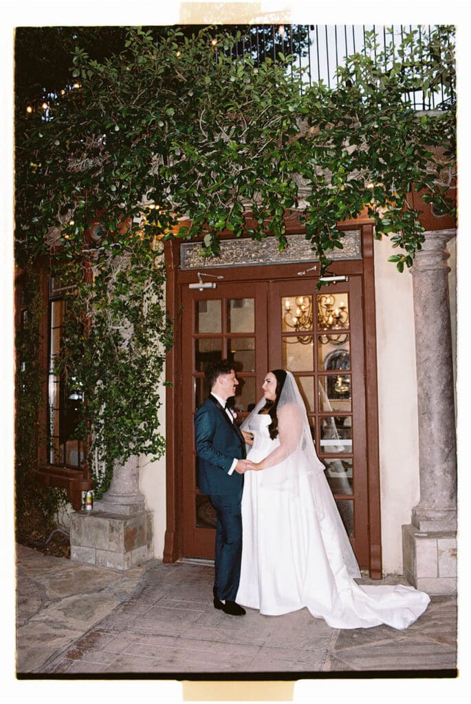 Bride and groom standing together beneath tall wooden doors framed by greenery and stone walls.