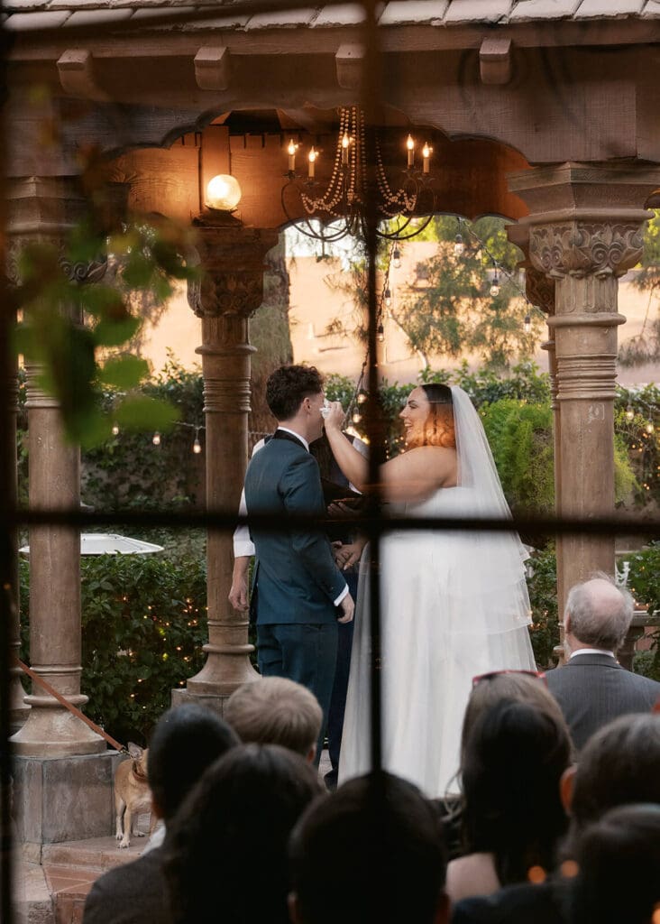 Wedding ceremony beneath an ornate wooden pavilion photographed through the seated guests.
