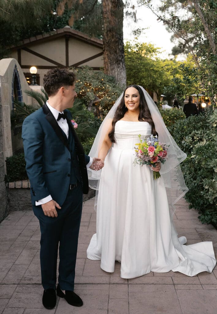 Bride and groom holding hands in a garden courtyard, with the bride carrying a colorful floral bouquet.